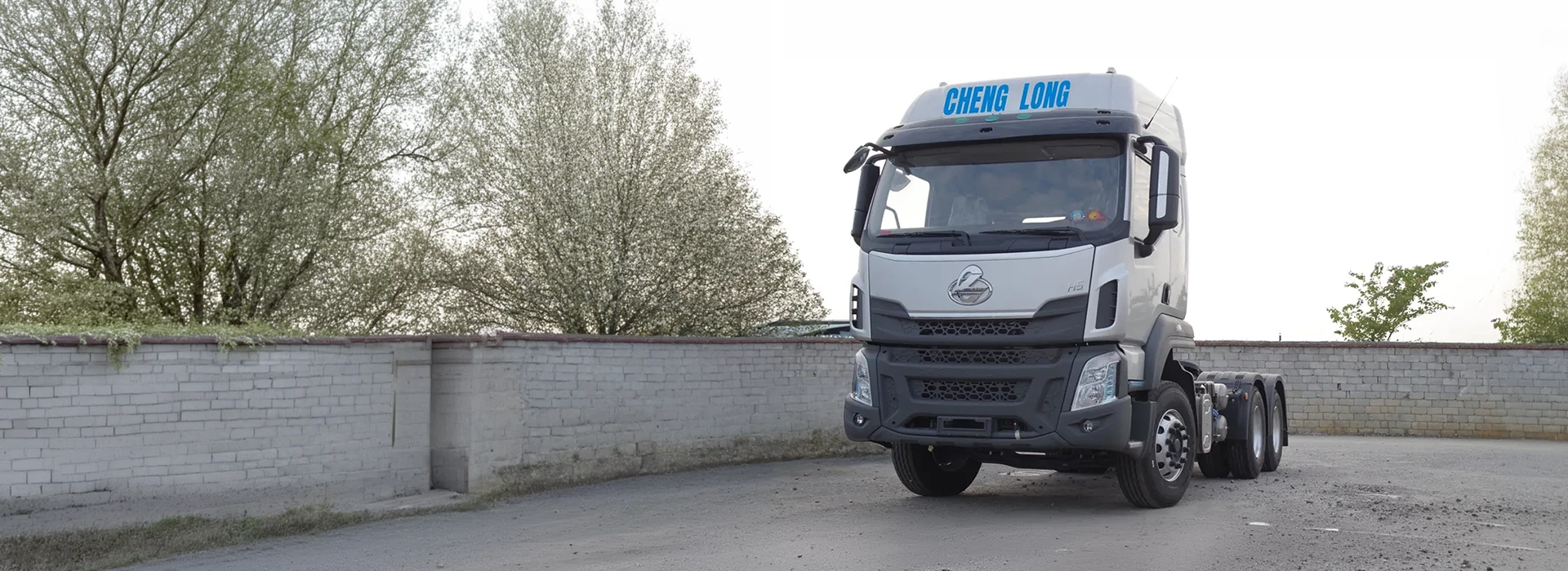 Silver Chenglong H5 6x4 tractor truck parked outdoors on an asphalt driveway next to a brick wall