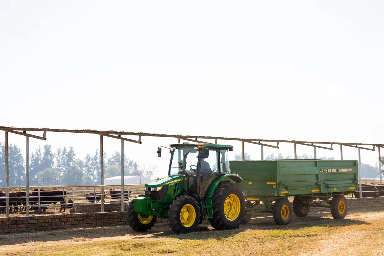 A green John Deere 5E Series cab tractor towing a trailer on a farm.