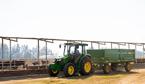 A green John Deere 5E Series cab tractor towing a trailer on a farm.