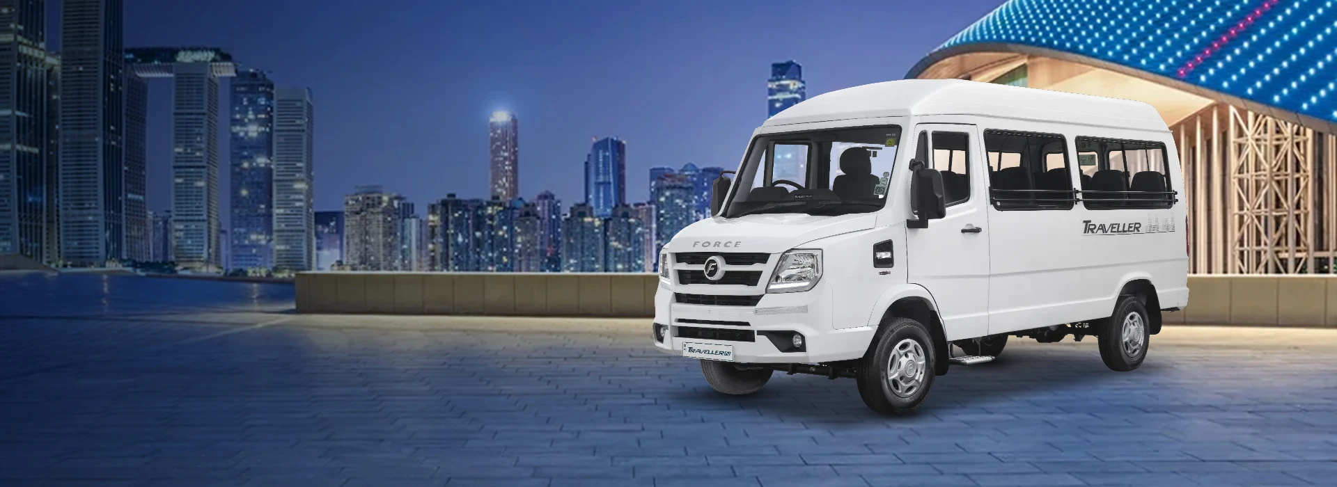 White Force Traveller high-roof passenger minibus parked on a paved plaza with an illuminated modern architectural building and urban skyline background at dusk