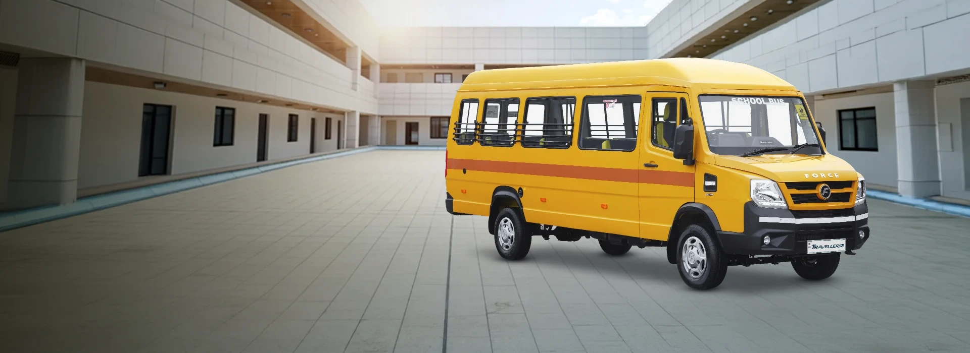 Yellow Force Traveller high-roof school bus with safety window guards and an orange side stripe, parked in front of a modern campus building