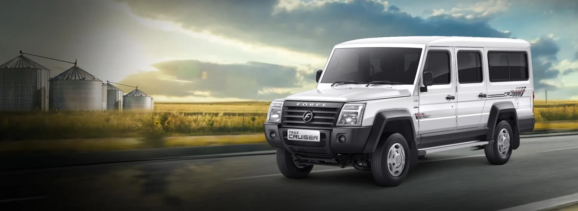 White Force Trax Cruiser multi-utility passenger vehicle driving on a paved road alongside agricultural fields and grain silos under a cloudy sky