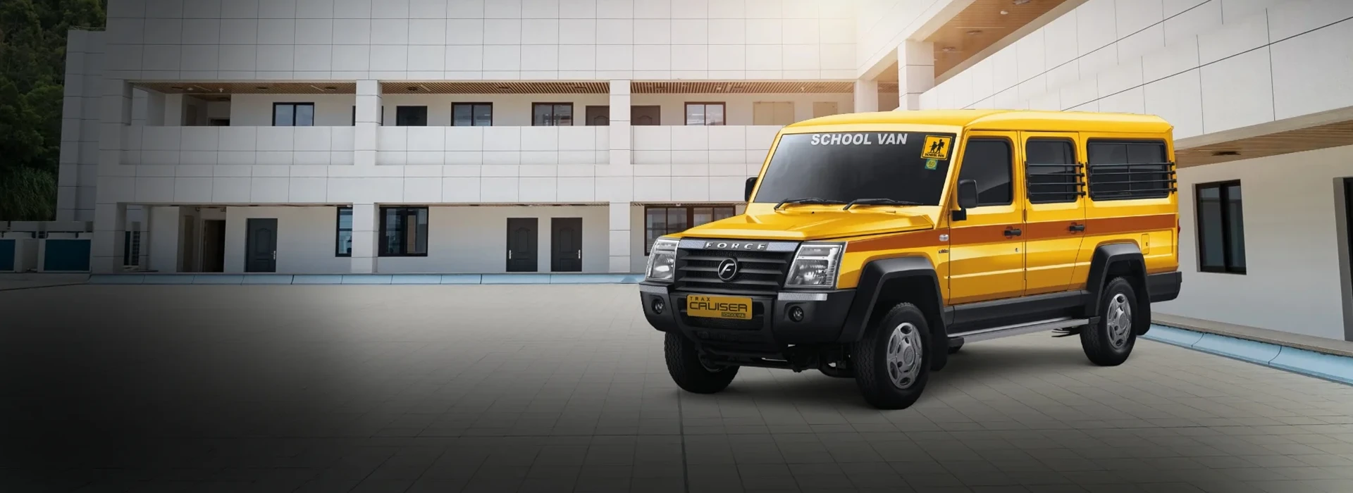 Yellow Force Trax Cruiser school van featuring safety window grills and an orange side stripe, parked in a paved campus courtyard in front of a modern building