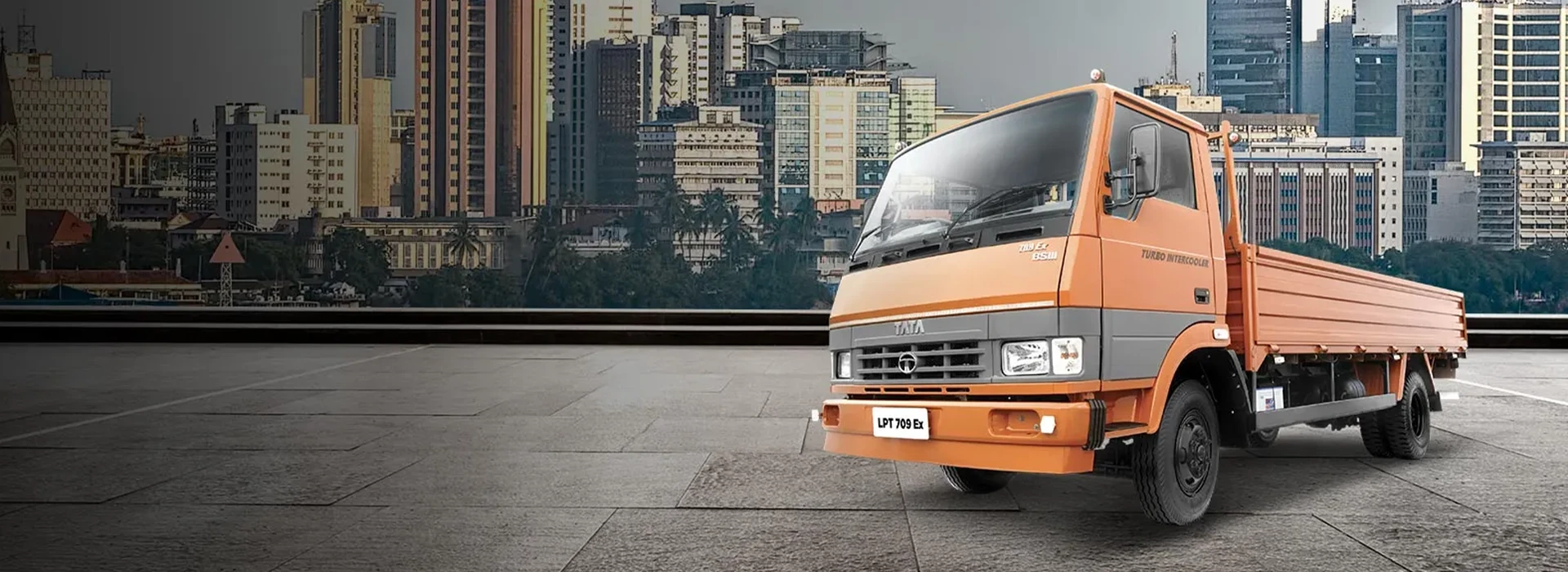 Orange Tata LPT 709 Ex flatbed truck parked on a paved platform with a city skyline in the background