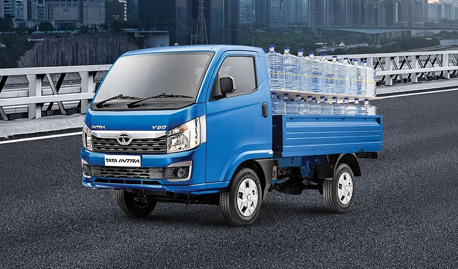Blue Tata Intra V20 pickup truck loaded with large water bottles on driving on a road with a city in the background.