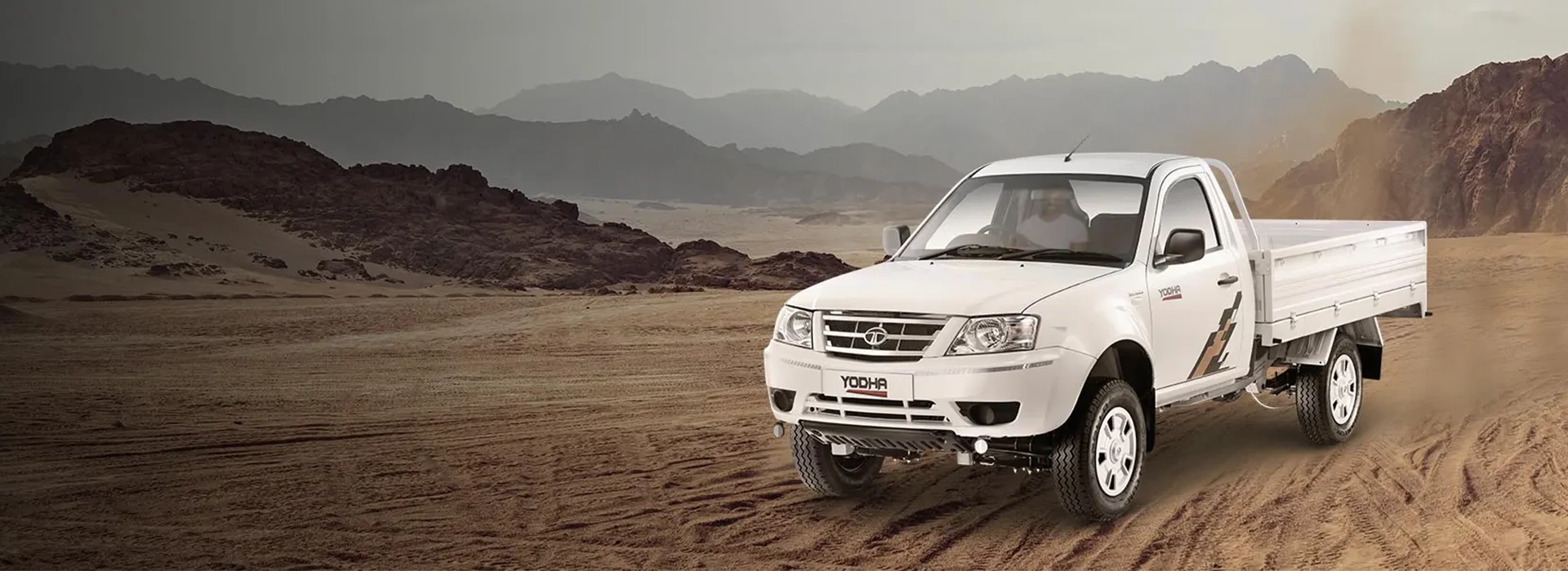 White Tata Yodha pickup truck driving on a sandy desert track with mountains in the background.