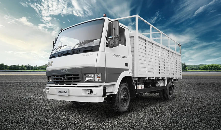 White and grey Tata LPT 810 Ex2 cargo truck with a drop-side body and overhead frame parked on an asphalt surface against a cloudy blue sky.