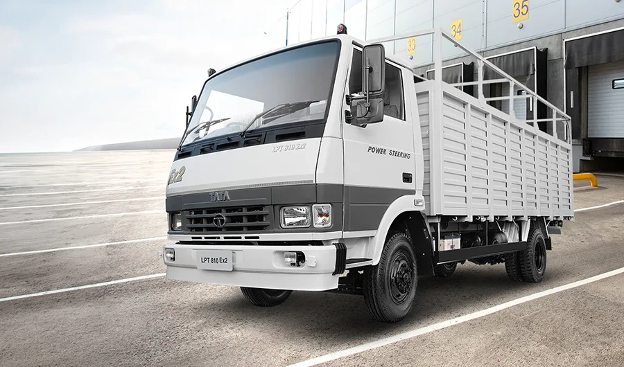 White and grey Tata LPT 810 Ex2 cargo truck with a drop-side body and overhead frame parked outside a warehouse loading dock.