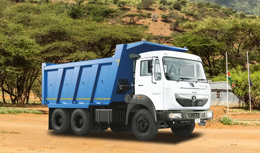 A white and blue Tata Signa heavy-duty tipper truck parked on a dirt terrain outdoors with sparse trees and a building in the background