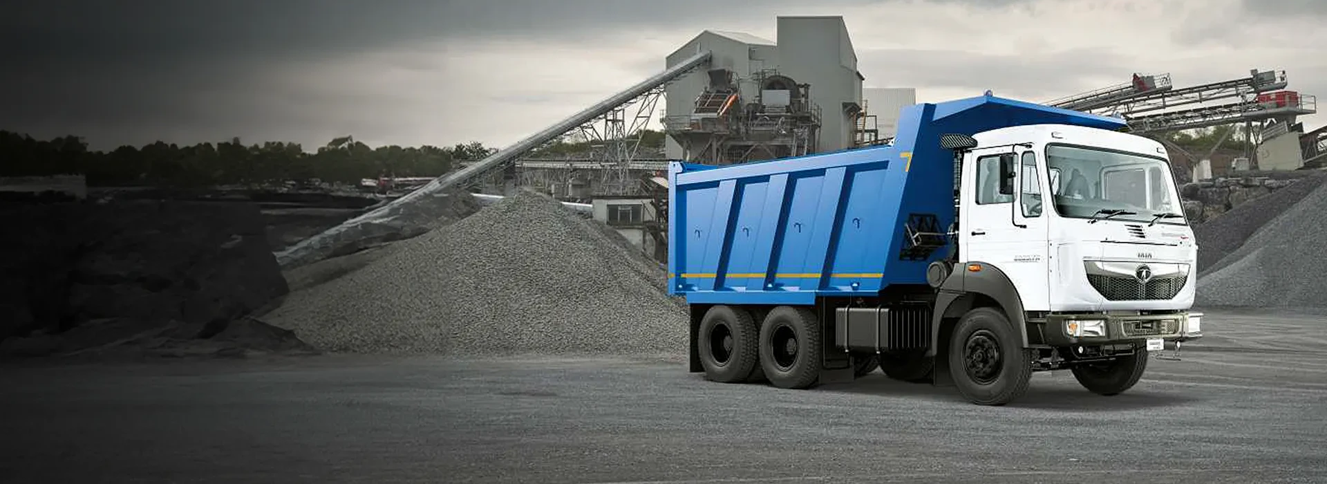 A white and blue Tata heavy-duty dump truck parked at a gravel quarry with industrial structures in the background.
