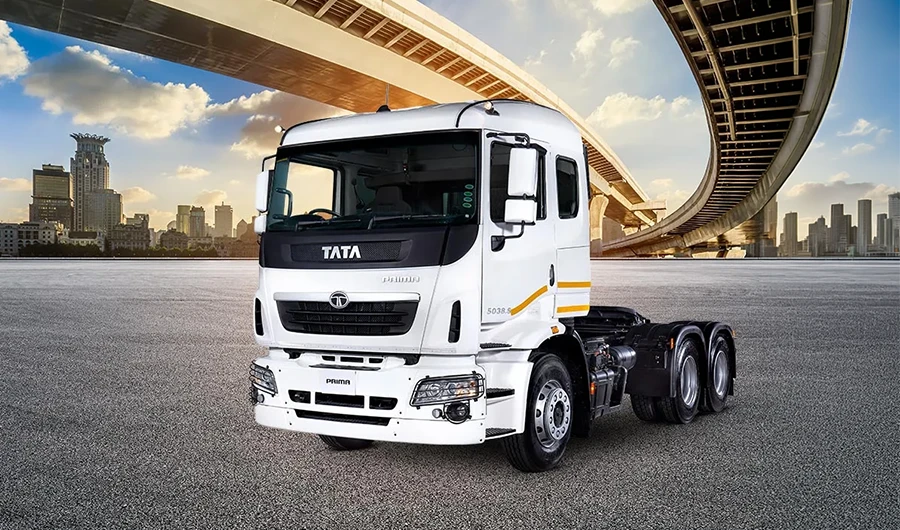 A white Tata Prima heavy-duty truck tractor parked on a paved surface with an urban cityscape and highway overpass in the background.