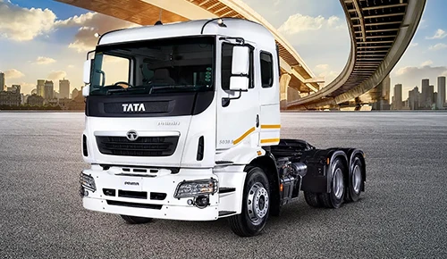 A white Tata Prima heavy-duty truck tractor parked on a paved surface with an urban cityscape and highway overpass in the background.