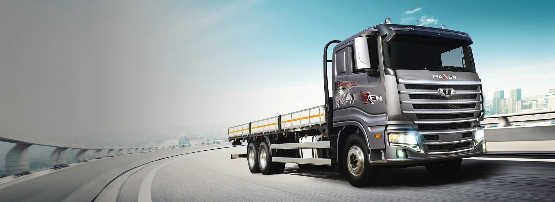 Metallic grey Daewoo Maxen heavy-duty flatbed truck driving along an elevated highway with headlights illuminated