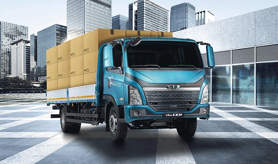 Teal blue Daewoo Dexen flatbed truck fully loaded with cardboard cargo boxes outside a modern building