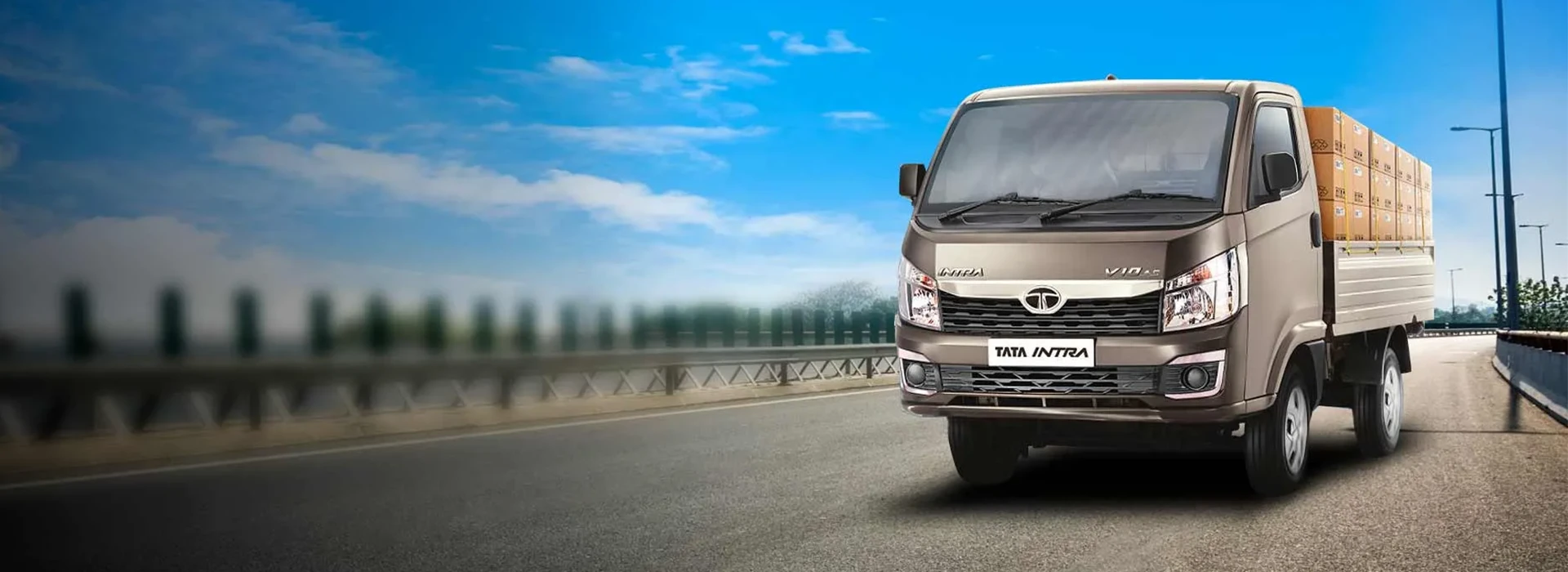 Front three-quarter view of a silver Tata Intra V10 light commercial truck carrying stacked cargo boxes on a highway against a blue sky background, styled as a wide hero banner