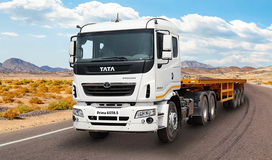 White Tata Prima 6038.S tractor head pulling a yellow flatbed trailer on a winding desert road