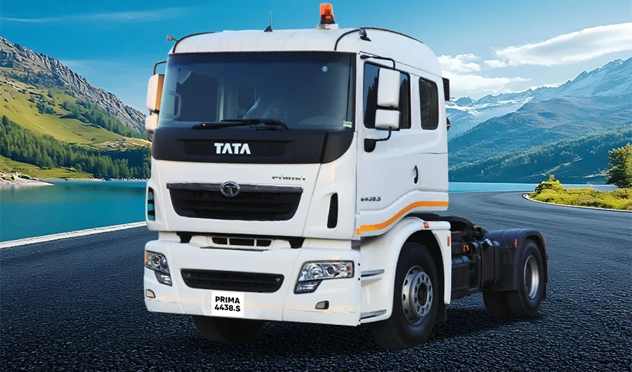 A white Tata Prima 4438.S truck tractor stationary on a paved road set against a backdrop of a mountain lake and alpine scenery under a clear blue sky.