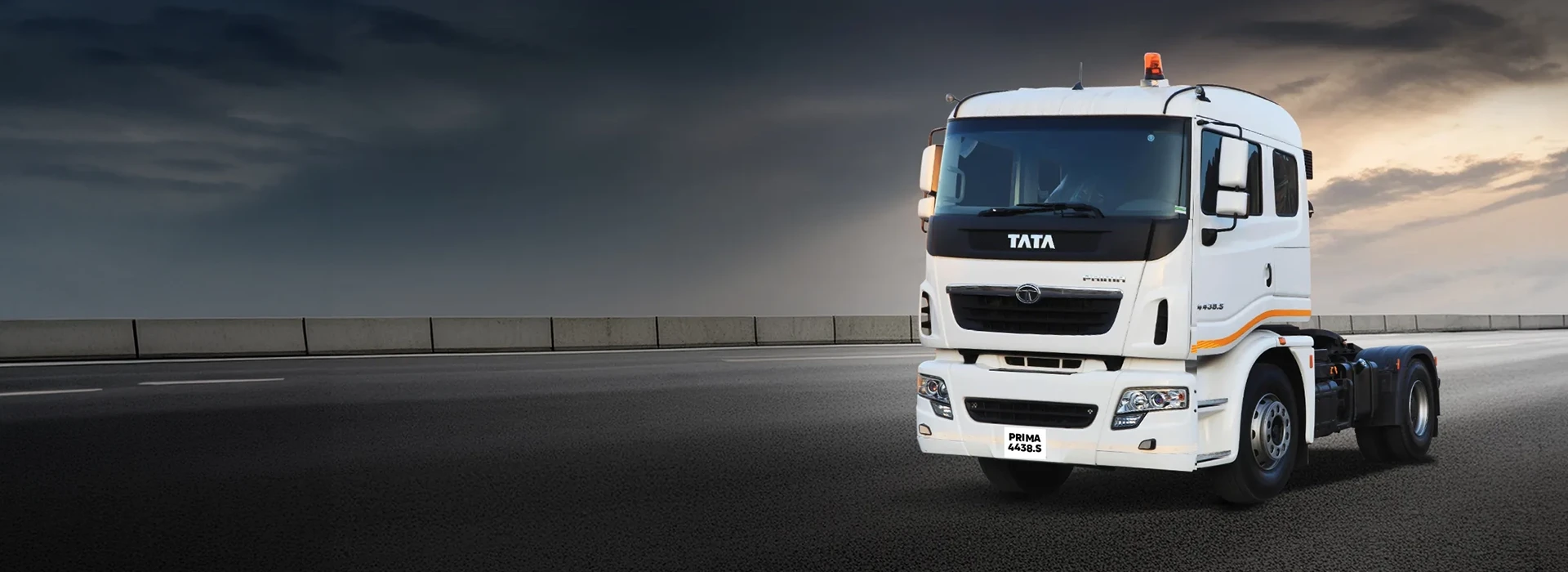 A front three-quarter view of a white Tata Prima 4438.S heavy-duty truck tractor driving on an open asphalt highway under a dramatic, overcast evening sky.