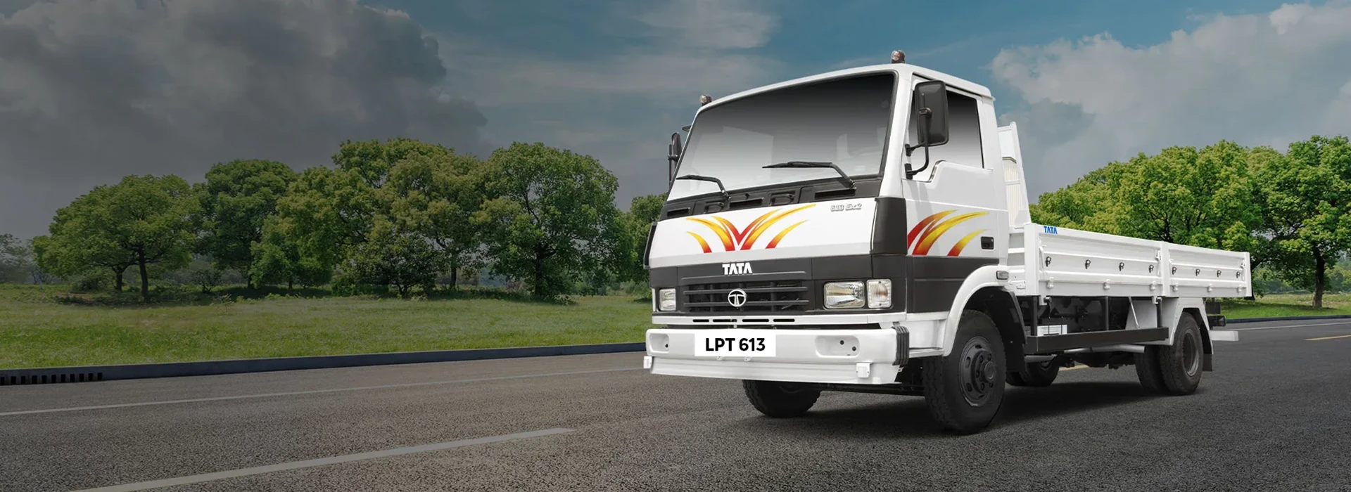 White Tata LPT 613 drop-side flatbed truck on an open paved road with trees and cloudy sky background
