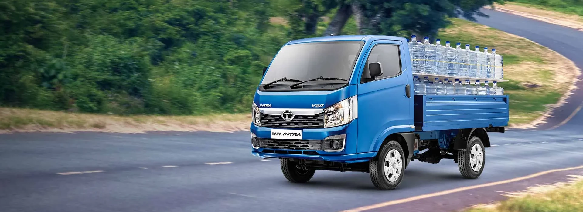 Blue Tata Intra V20 pickup truck loaded with large water bottles driving on a winding scenic road.