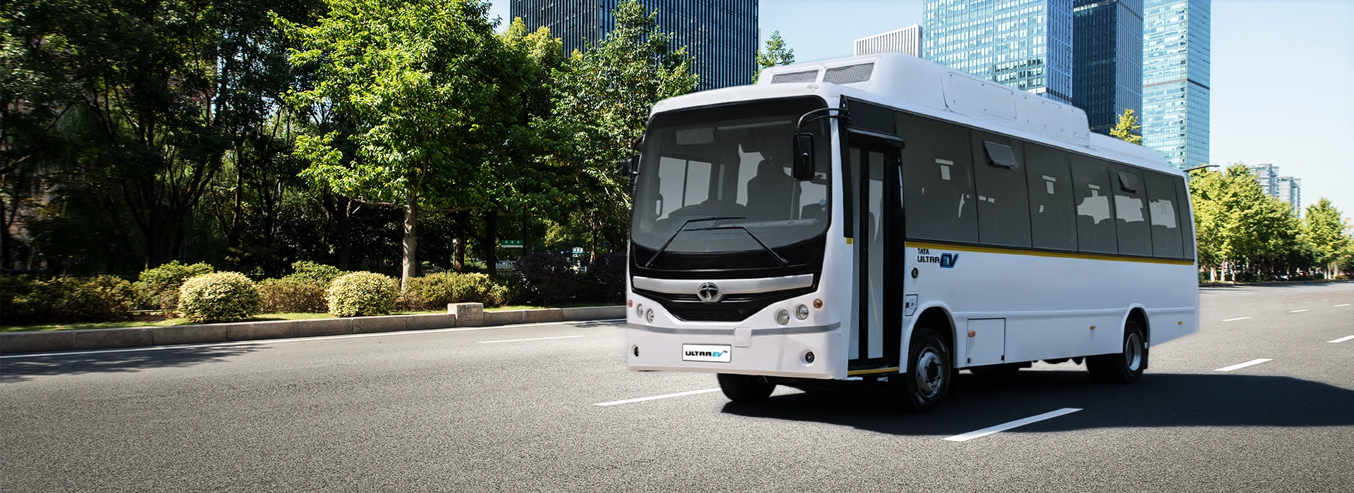 White Tata Ultra passenger bus driving on a city main road with trees and tall buildings in the background