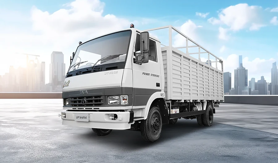 White and grey Tata LPT 810 Ex2 cargo truck with a drop-side body and overhead frame parked on a paved surface with a city skyline background.