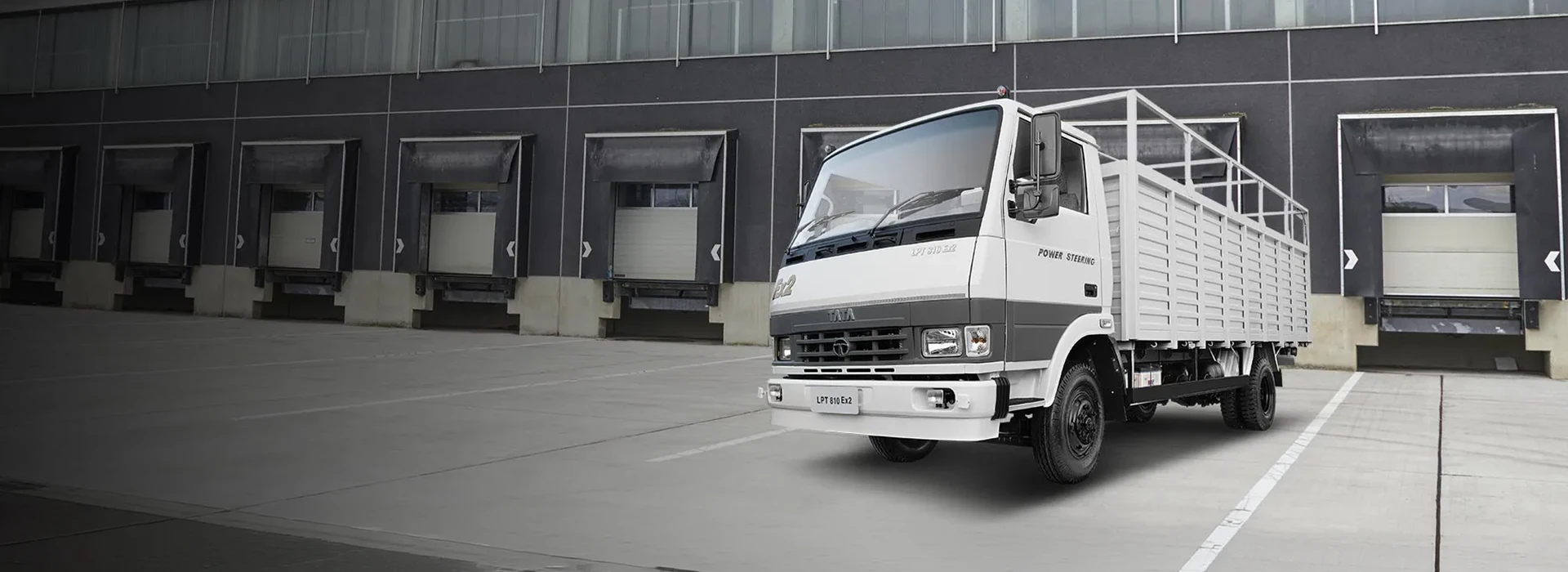 White and grey Tata LPT 810 Ex2 cargo truck with a drop-side body and overhead frame parked outside an industrial warehouse.