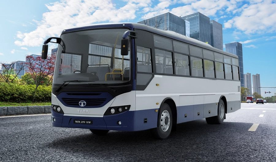 A three-quarter front-side view of a blue and white Tata LPO 1318 commuter coach bus on a paved city road, with modern glass-fronted office buildings and green landscaping in the background under a partly cloudy sky.