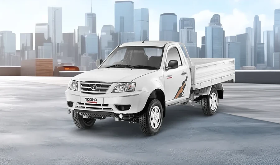 White Tata Yodha pickup truck parked on a paved surface with a city skyline background.