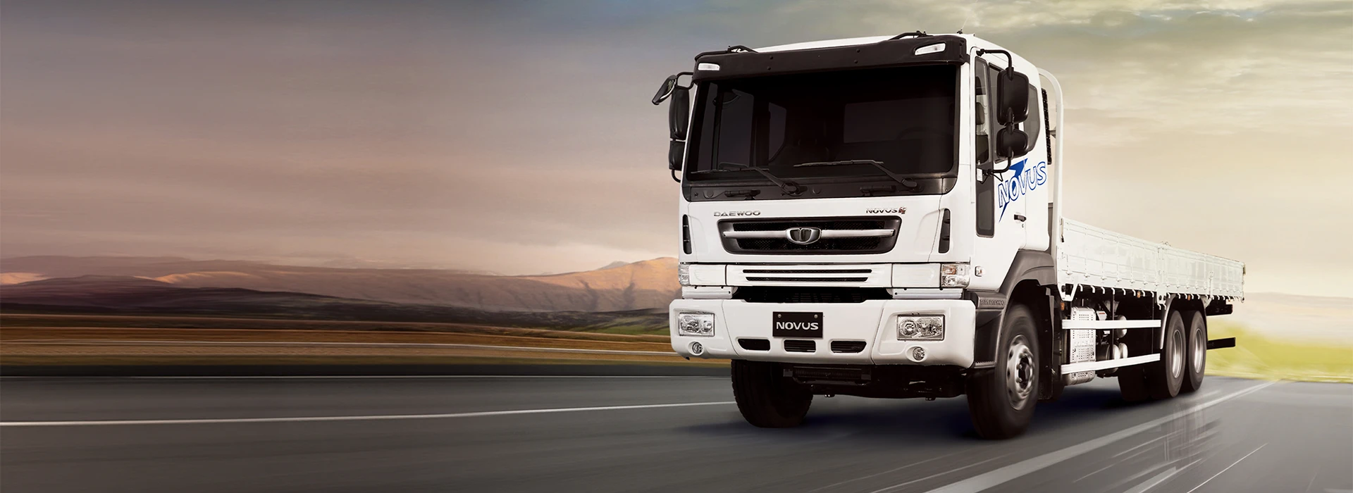 White Daewoo Novus flatbed truck driving along an open highway at sunset