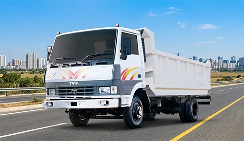 A front three-quarter view of a white Tata LPK 813 Tipper truck driving on a asphalt highway, set against a background of a modern city skyline under a bright blue sky.