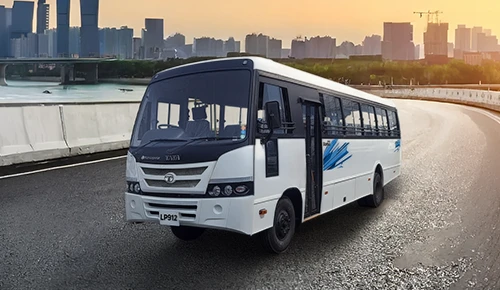 White Tata LP 912 passenger bus driving on an open road with a city skyline at sunset in the background